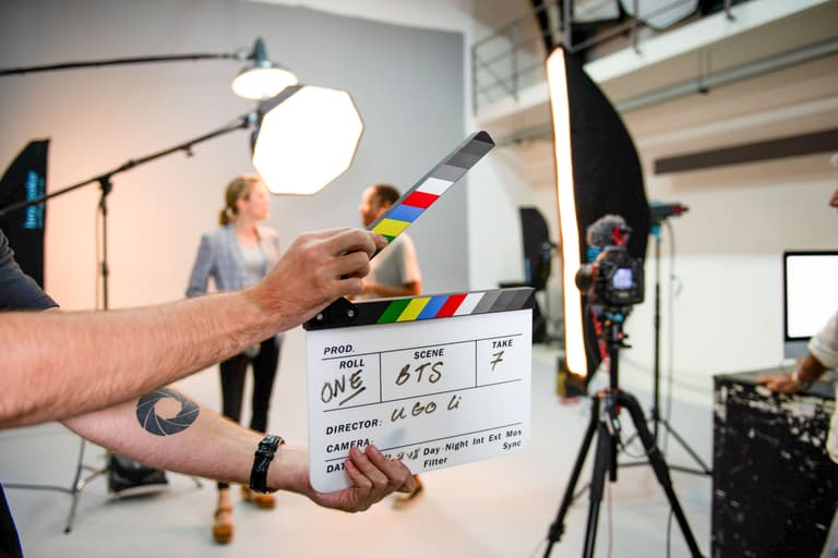 A clapperboard is being held on a busy film set with bright lighting and crew.
